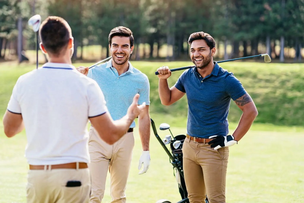 Three men enjoying a conversation on a golf course, with golf clubs in hand and golf bags nearby.