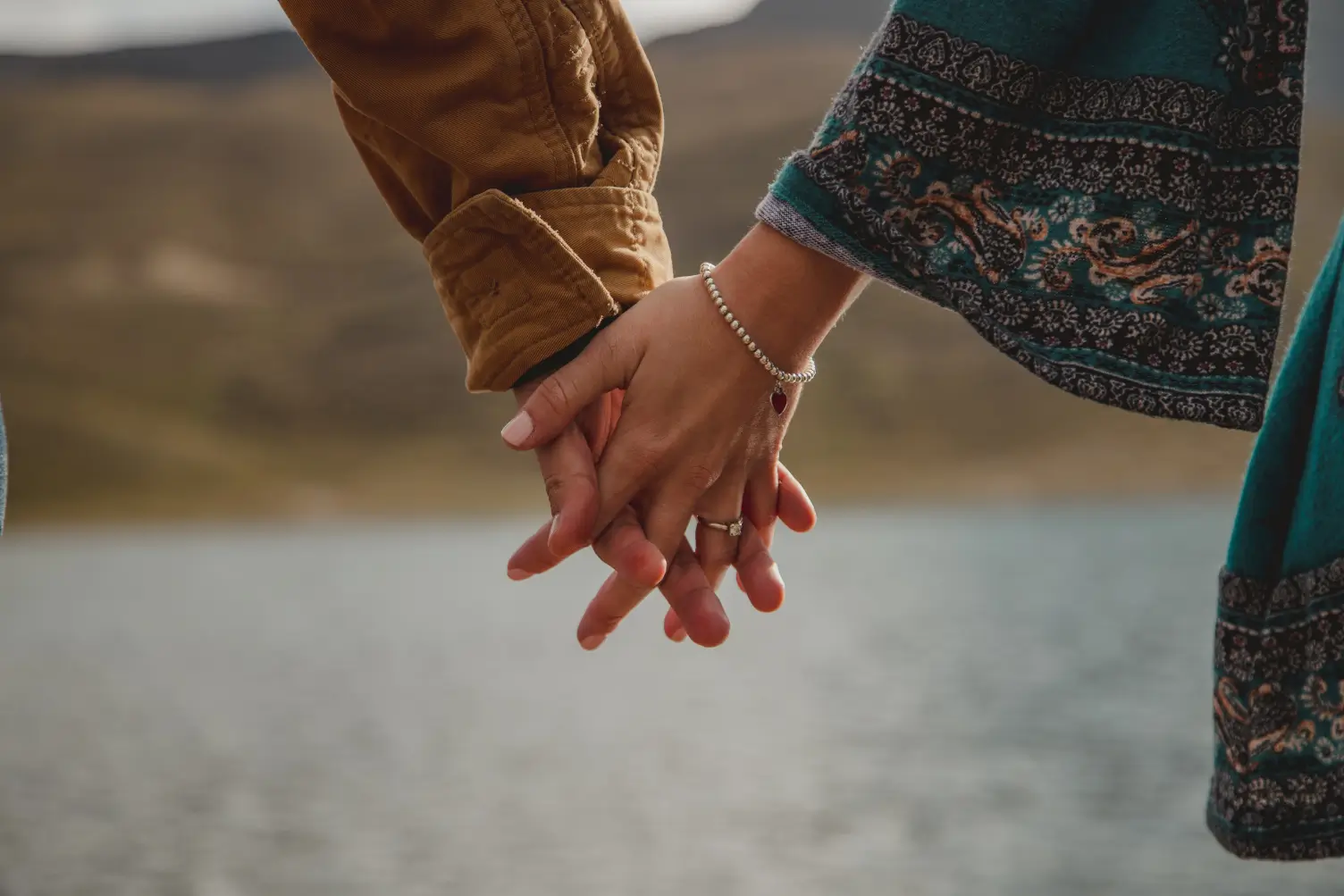 A close-up of two hands holding each other in front of a tranquil water body, symbolizing connection and intimacy in a relationship after 90 days of relationship coaching to transform their relationship.