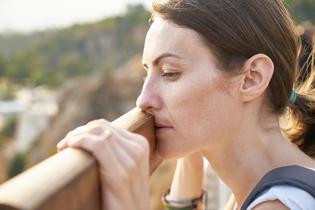 A woman with a pensive expression resting her chin on a wooden railing in a natural setting.