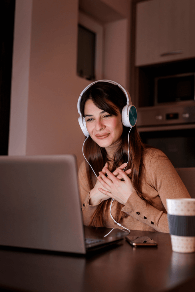 A relationship coach online wearing headphones sits at a desk, smiling while looking at a laptop screen, with a coffee cup and a phone nearby.