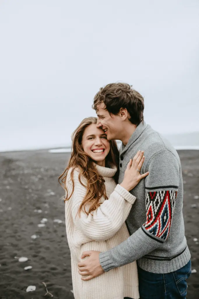 A couple smiling and embracing each other outdoors on a sandy beach, dressed in cozy sweaters, expressing joy and connection after relationship coaching for women in NJ.